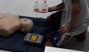 A person is kneeling beside a medical training dummy, holding defibrillator paddles connected to a defibrillator machine, practicing CPR on a dark mat. The person is wearing a white shirt and dark shorts.