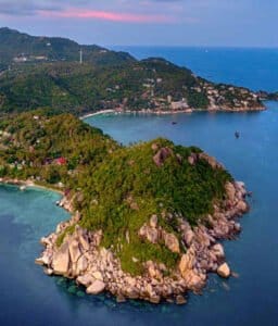 An aerial view of Koh Tao, revealing lush green hillsides meeting pristine white sand beaches and a calm, turquoise ocean dotted with traditional long-tail boats.
