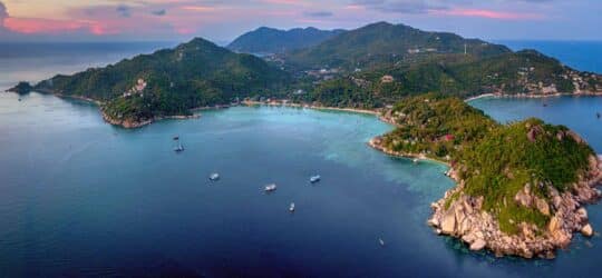 An aerial view of Koh Tao, revealing lush green hillsides meeting pristine white sand beaches and a calm, turquoise ocean dotted with traditional long-tail boats.